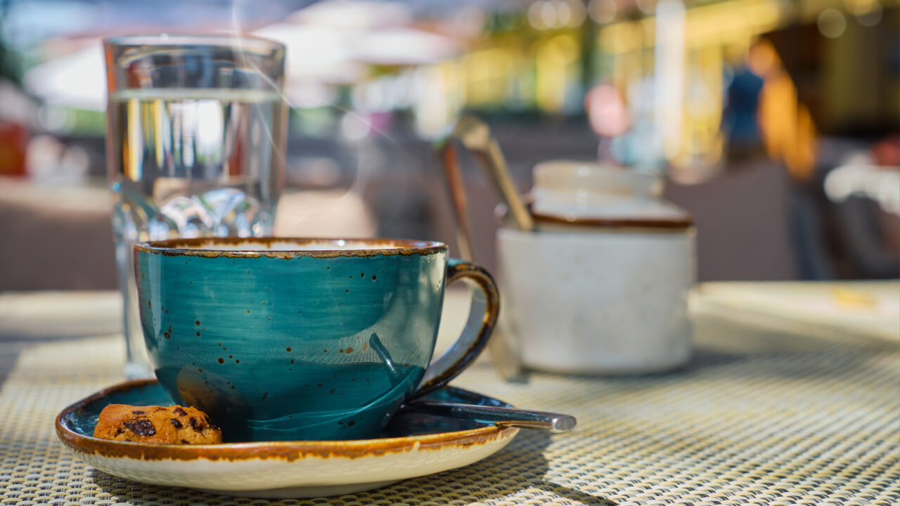 Steam rises over a cup of espresso coffee, a glass of water and cookies on the table, close-up, sunlight and selective focus, empty space, cafe breakfast, advertising or banner